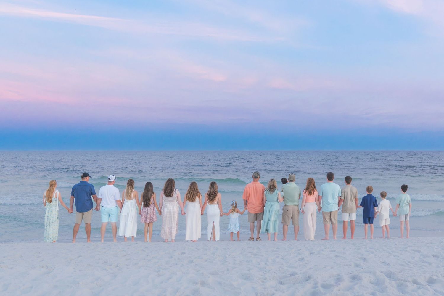 Large family group standing hand-in-hand on a beach at sunset with pastel pink and blue sky over calm ocean.