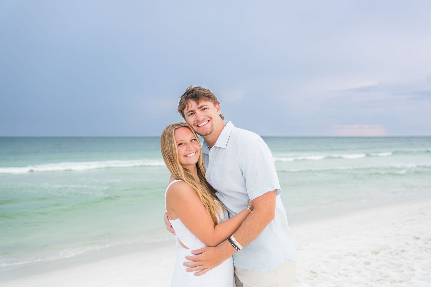 A smiling couple embraces on a white sandy beach with turquoise ocean waves and pastel sky behind them.