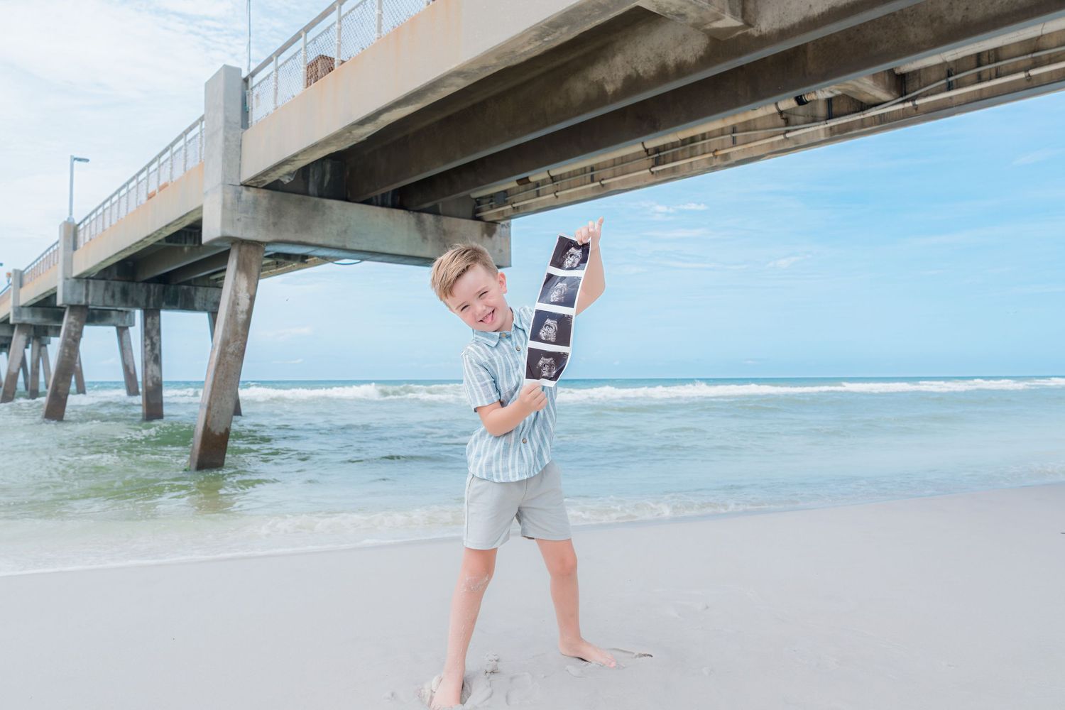 A young boy stands on a beach near a pier, proudly holding ultrasound photos, announcing a pregnancy.
