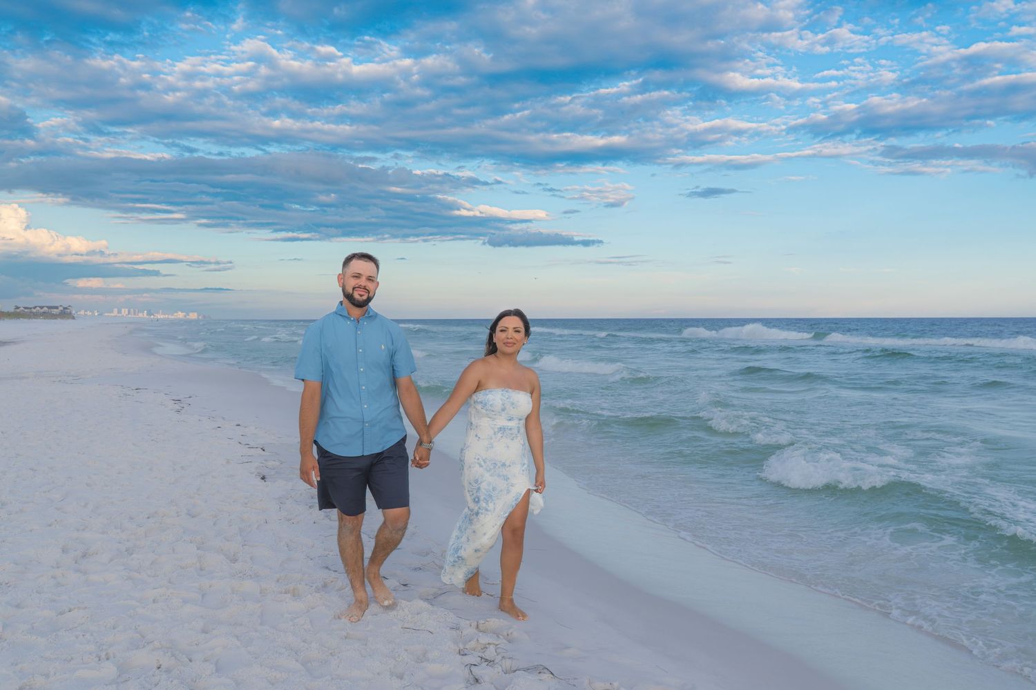 Couple holds hands walking along white sand beach at sunset with turquoise Gulf waters behind them.