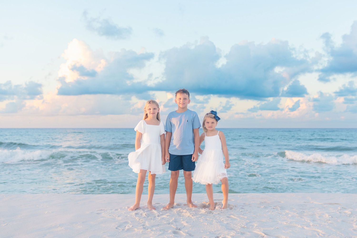 Three children pose on a white sand beach with turquoise waves and pastel clouds at sunset.