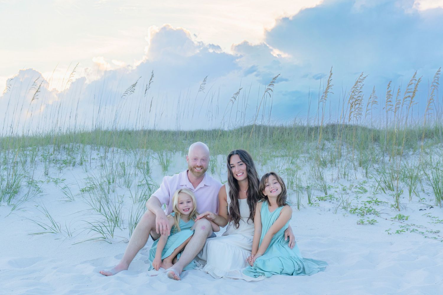 Family of four sitting together on white sand dunes with sea oats at beach sunset.
