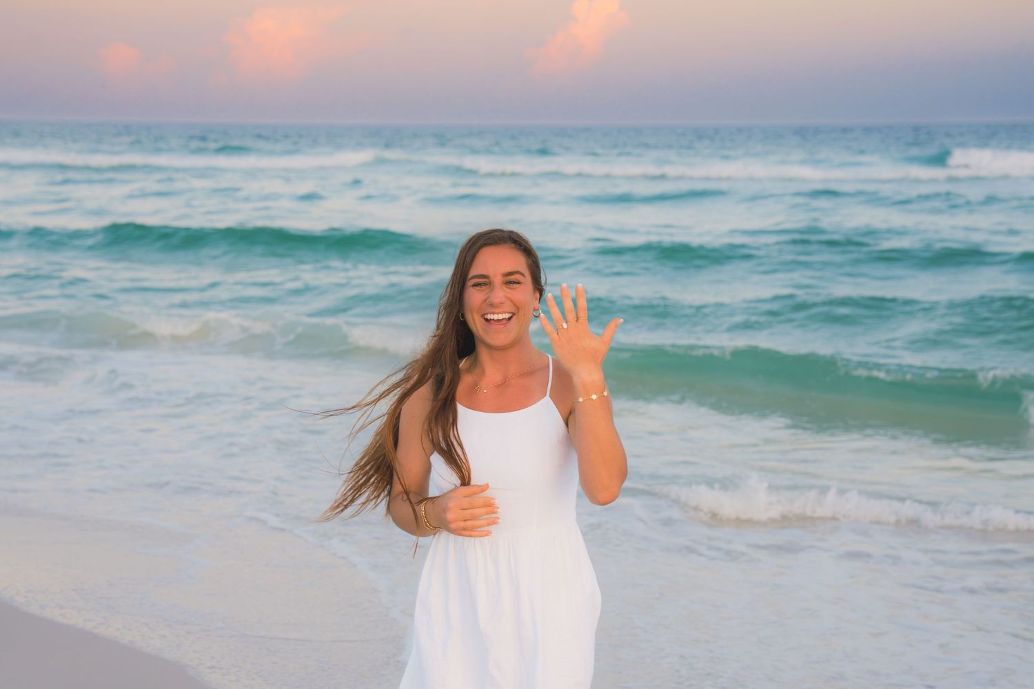 Woman in white dress waving and smiling on a tropical beach with turquoise waves at sunset.