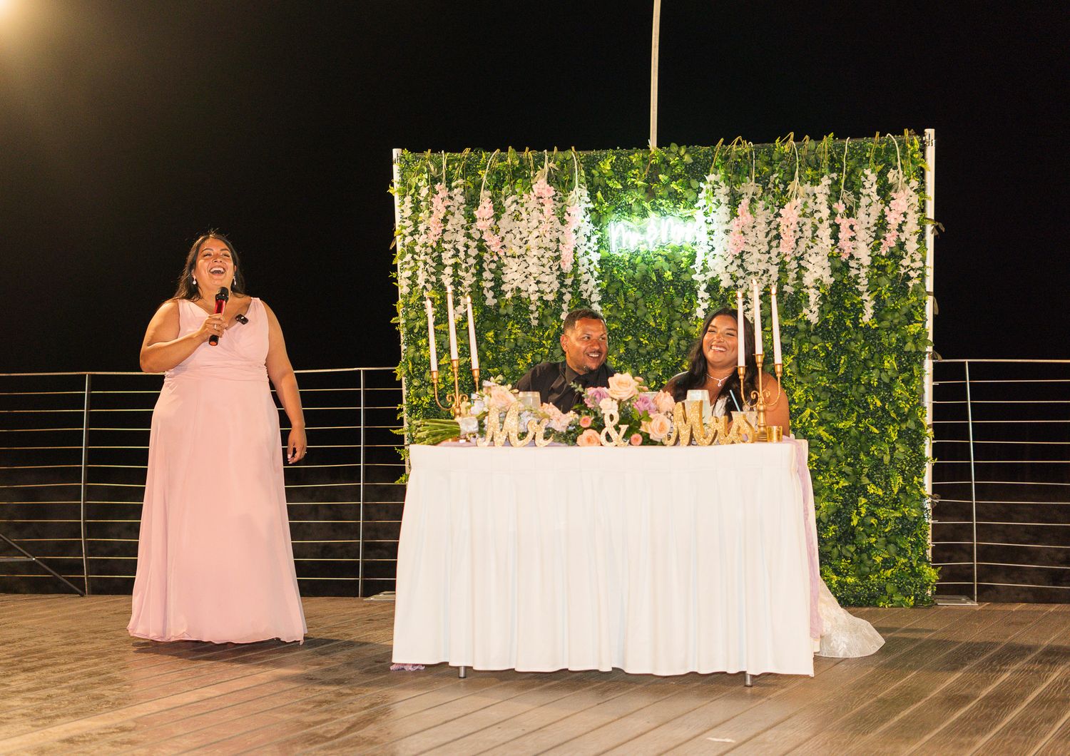 Bridesmaid gives a heartfelt speech as bride and groom sit at their floral sweetheart table at night.