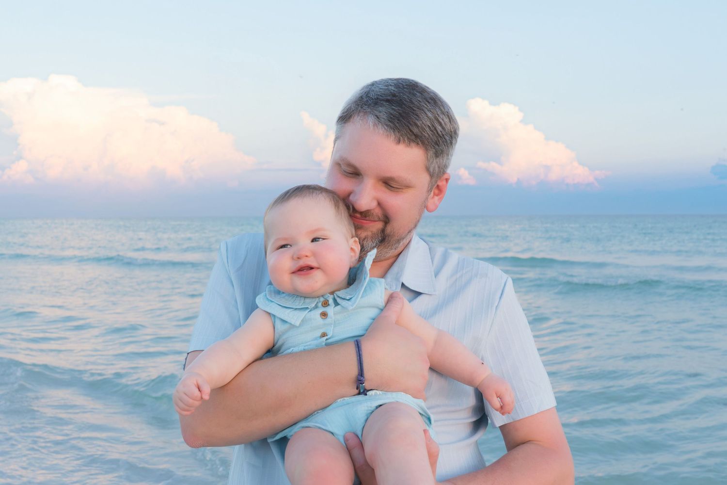 A father holds his smiling baby on a beach at sunset with calm ocean waves and pink clouds in the background.