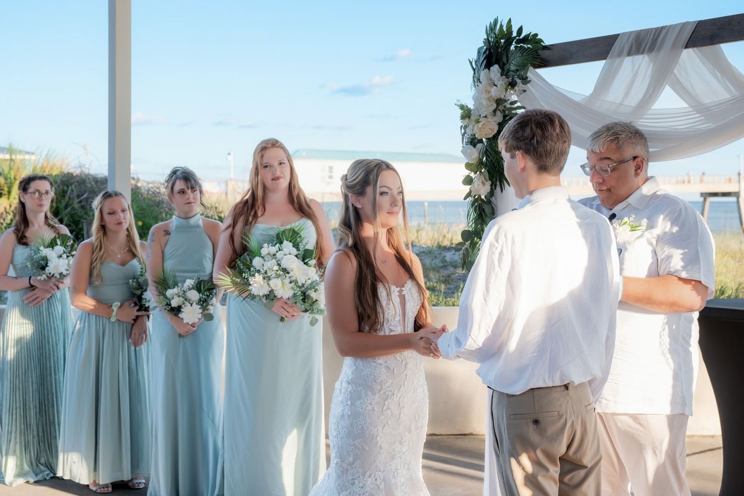 Bride in lace gown holds hands with groom at outdoor beach wedding ceremony with bridesmaids in blue dresses.