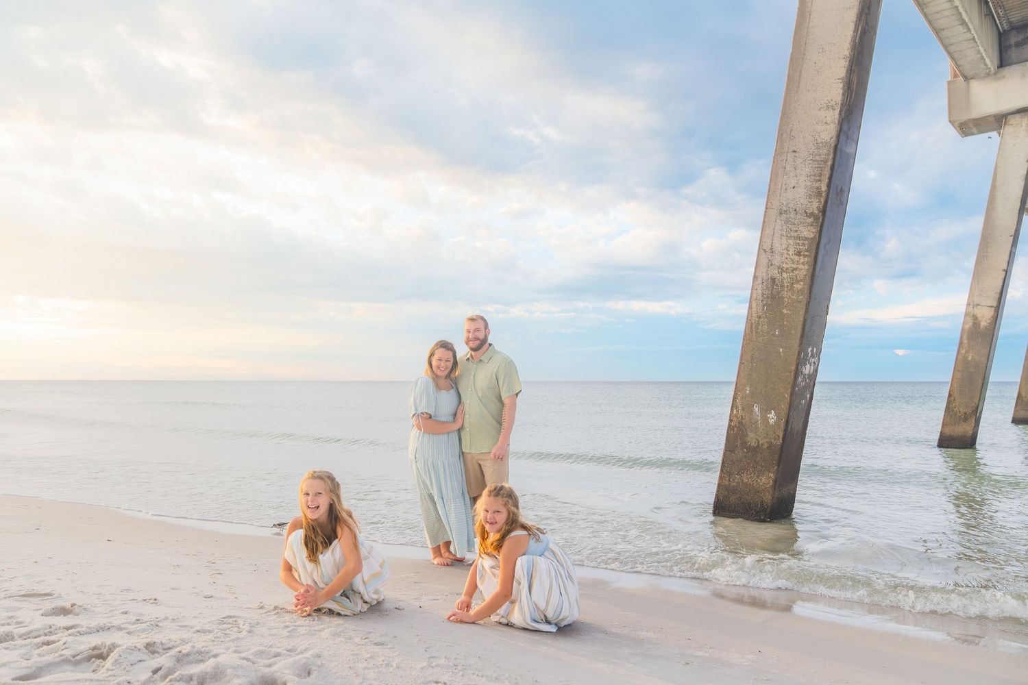 Family of four posing together on sandy beach near pier at sunset.