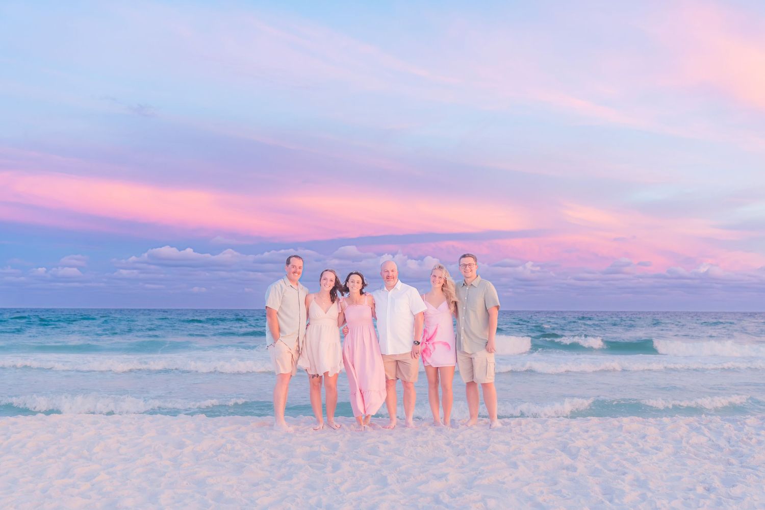 Family of six stands together smiling on a beach during a colorful pink sunset.