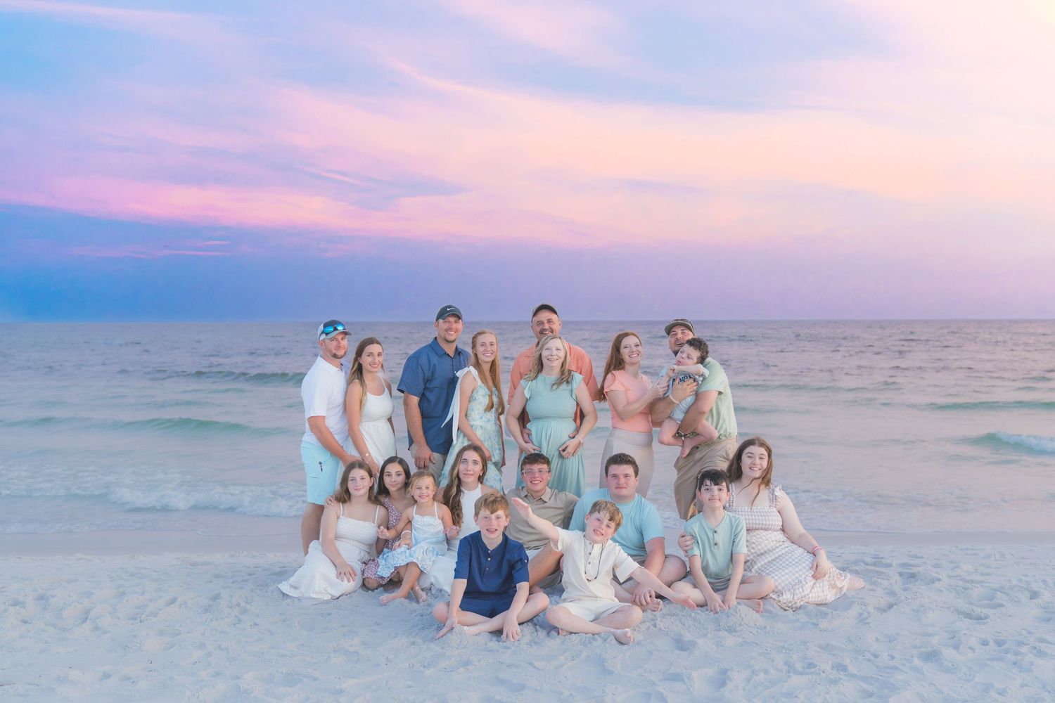Large family group photo on a beach at sunset with pastel pink and blue sky, wearing coordinated white and teal outfits.