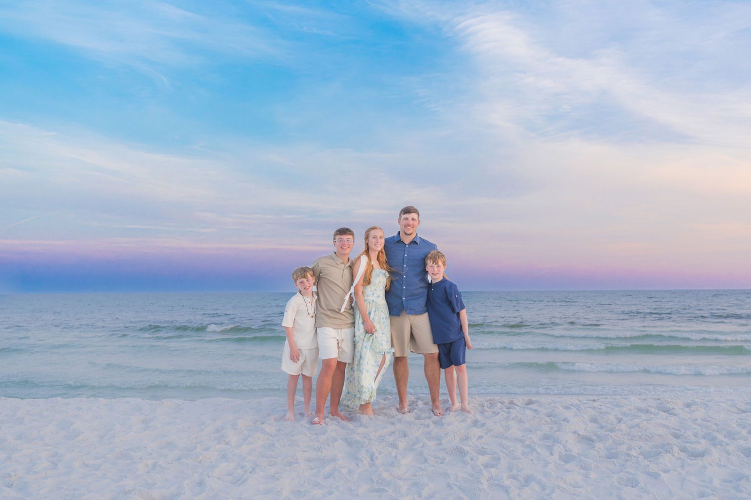 Family of five posing on a white sand beach at sunset with pastel pink and blue sky over calm ocean waters.