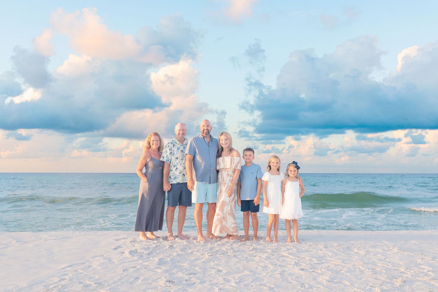Family of seven posing on a white sand beach at sunset with pastel clouds and turquoise ocean waves behind them.