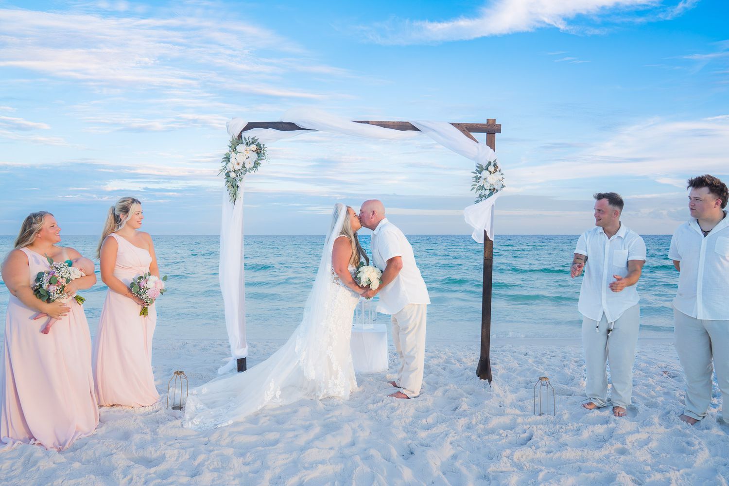 Bride and groom share first kiss at beach wedding ceremony under floral arch with bridesmaids in pink.