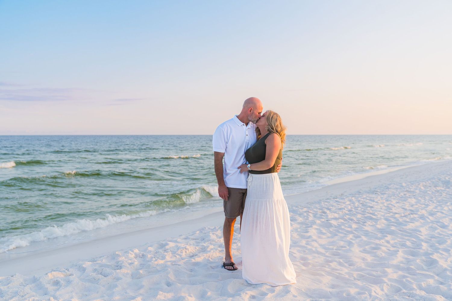 Couple sharing a romantic kiss on a white sand beach at sunset with calm turquoise ocean waves in the background.