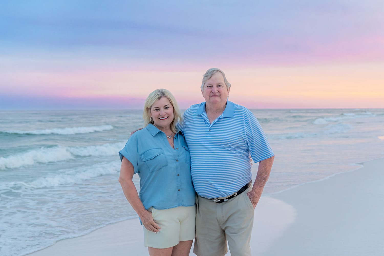 Couple smiling together on a sandy beach at sunset with pink and purple sky over calm ocean waves.