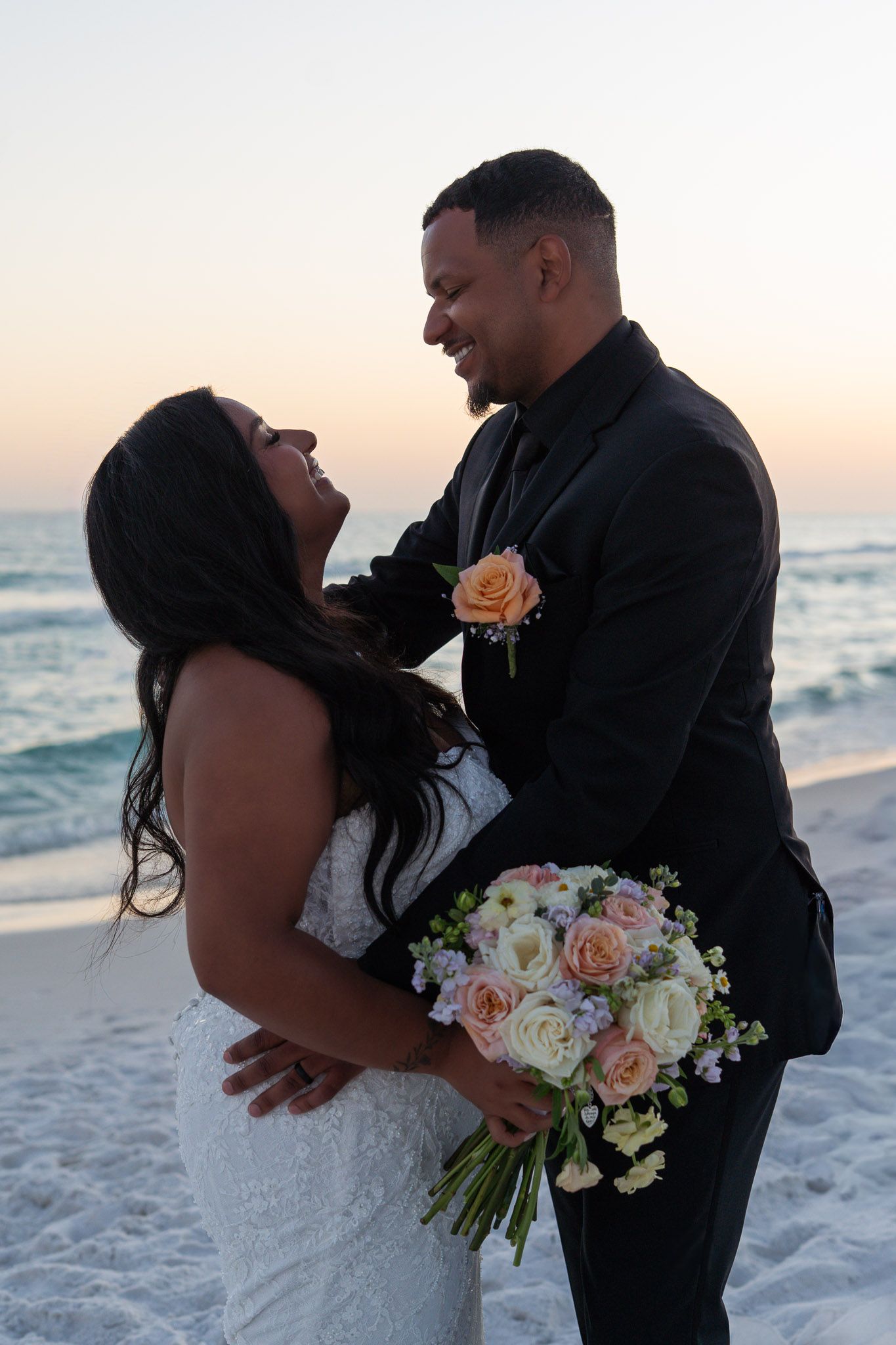 Newlywed couple embracing on a beach at sunset, bride holding a bouquet of pink and white roses in a lace wedding gown.