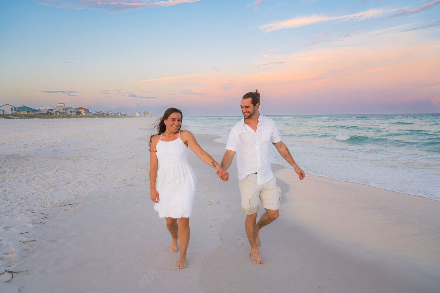 Couple in white outfits holding hands walking on a sandy beach at sunset with calm ocean waves.