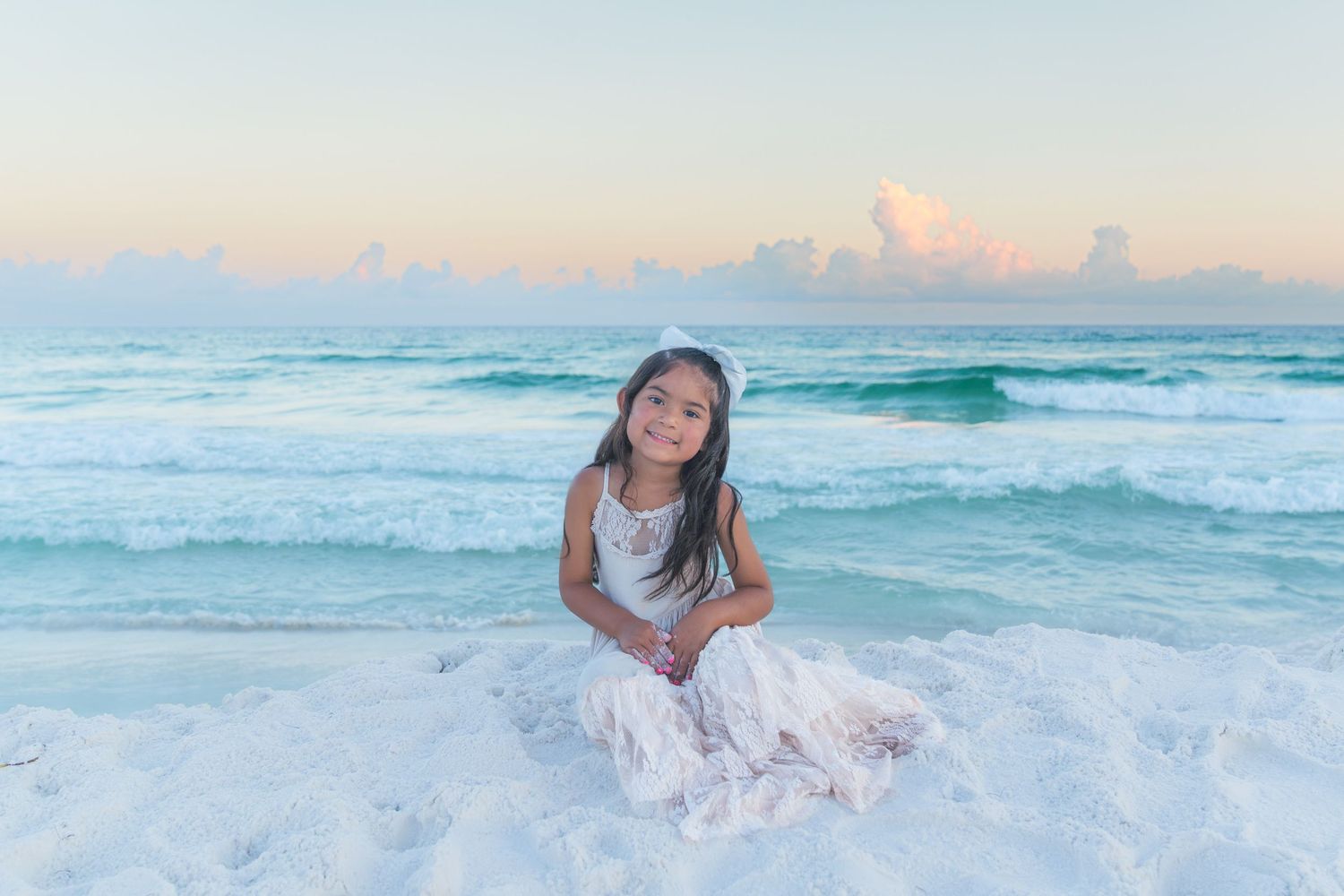 Young girl in white dress sitting on white sand beach with turquoise ocean waves and pastel sunset sky.