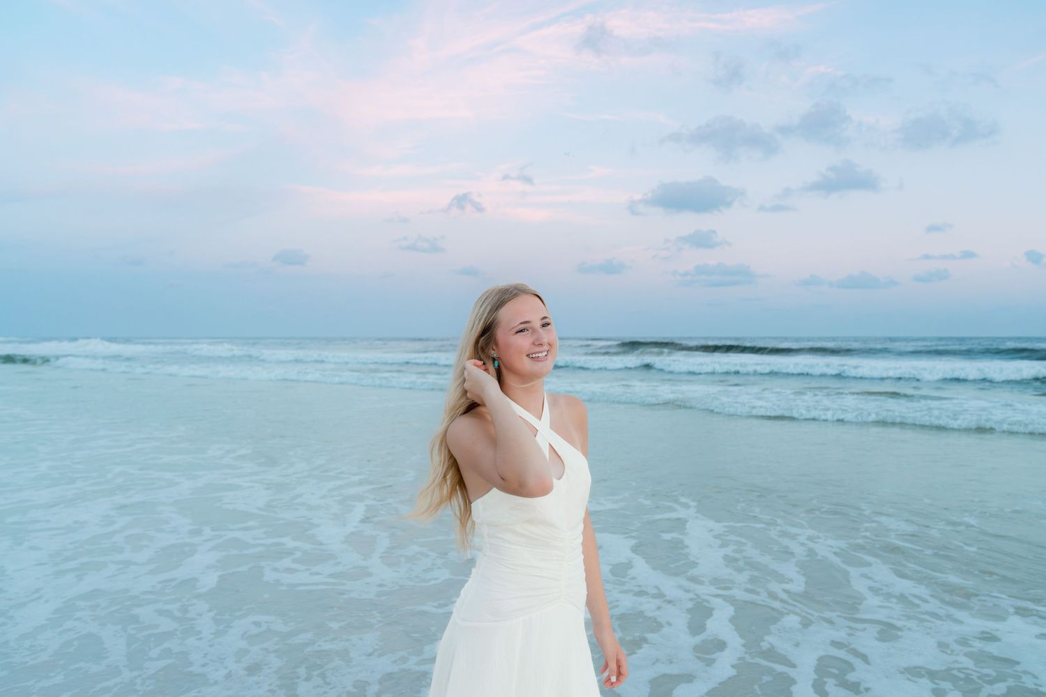 Young woman in white dress smiling on a beach at sunset with gentle waves and pastel sky.