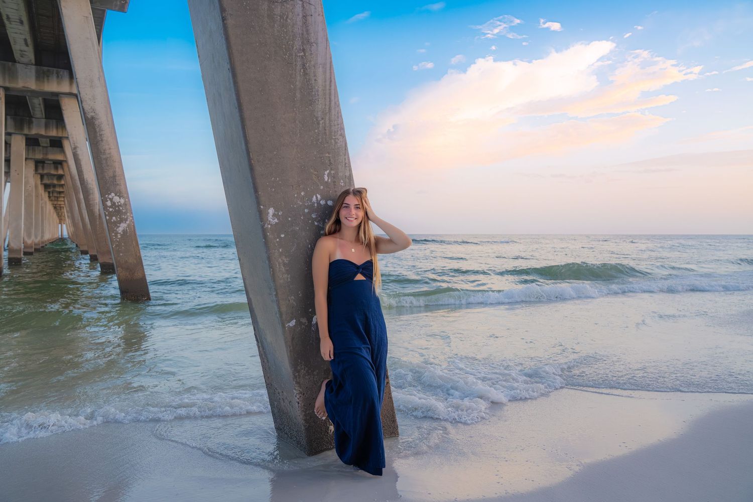 Woman in navy blue gown poses by beach pier pillars at sunset with ocean waves.