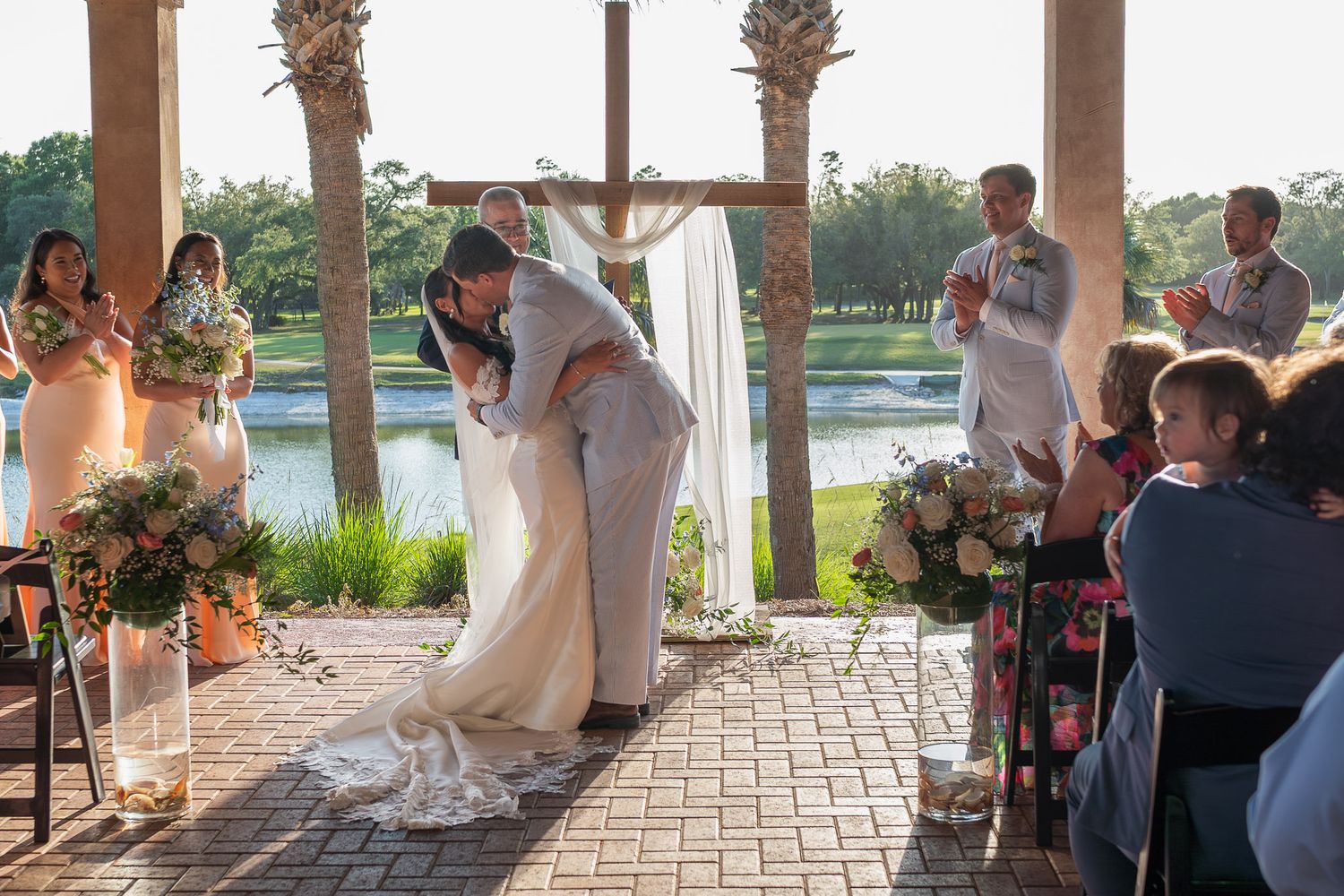 Bride and groom share first kiss at outdoor wedding ceremony by lakeside pavilion with guests applauding.