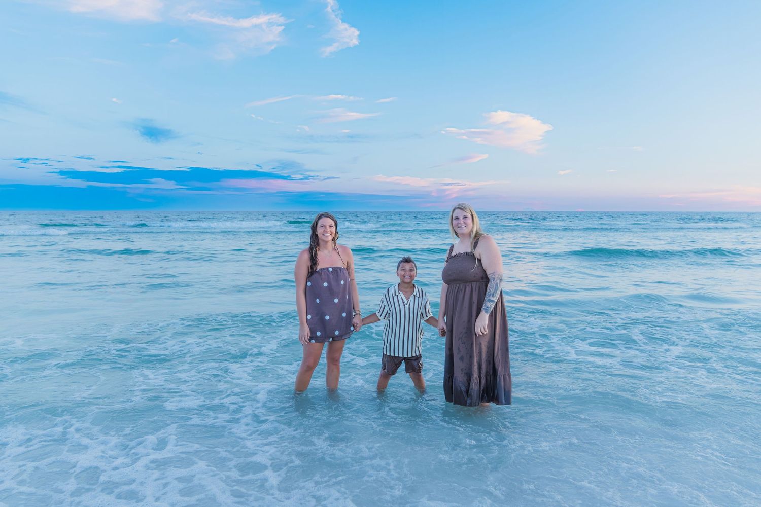 Two women and a child stand holding hands in shallow ocean water at sunset on a beautiful beach.