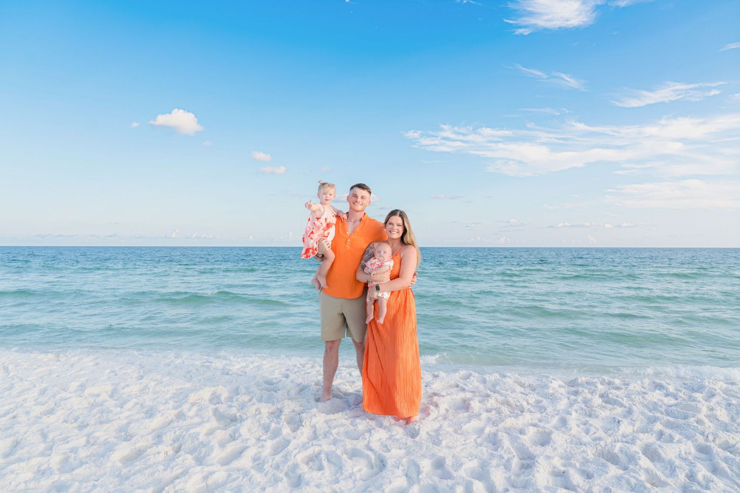 Family of three in matching orange outfits posing on a white sand beach with turquoise ocean water.