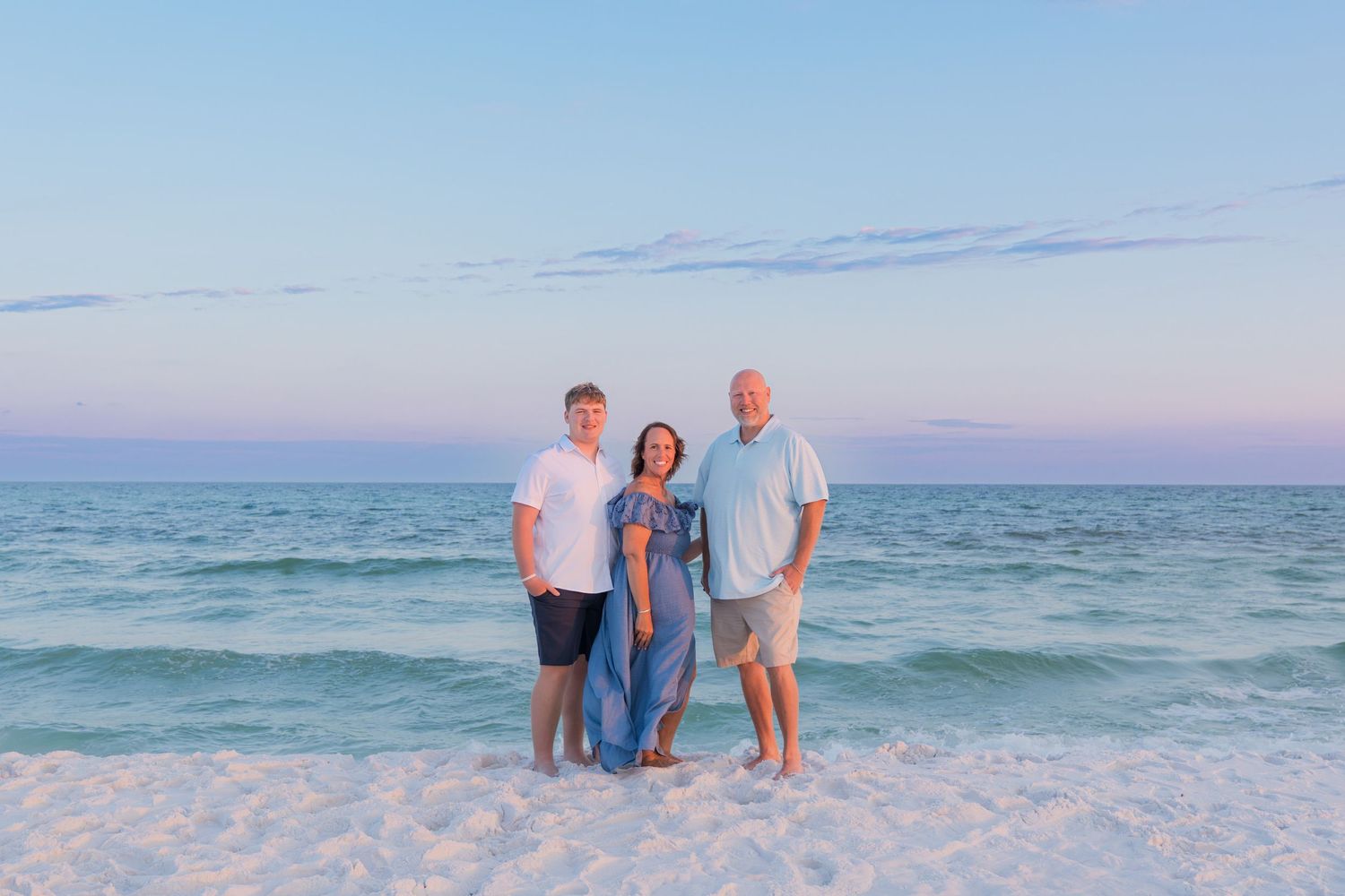 Three people smiling together on a white sand beach at sunset with turquoise ocean waves behind them.
