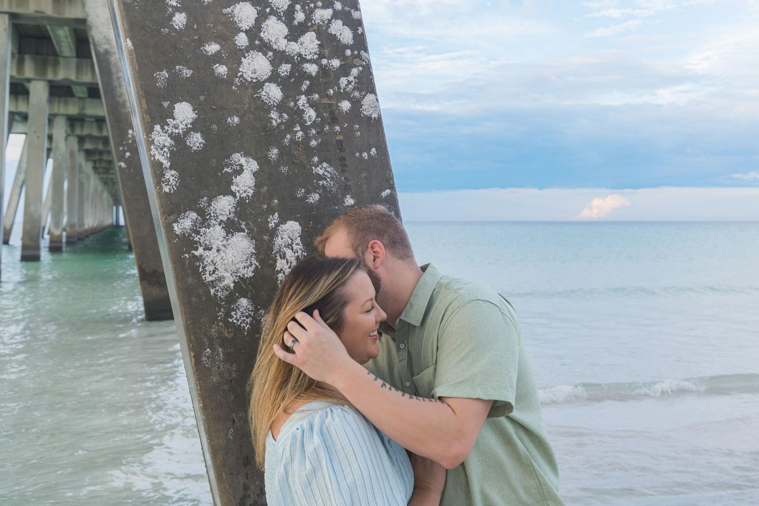 Couple sharing a romantic kiss leaning against a weathered pier piling on a beach with turquoise ocean waves.