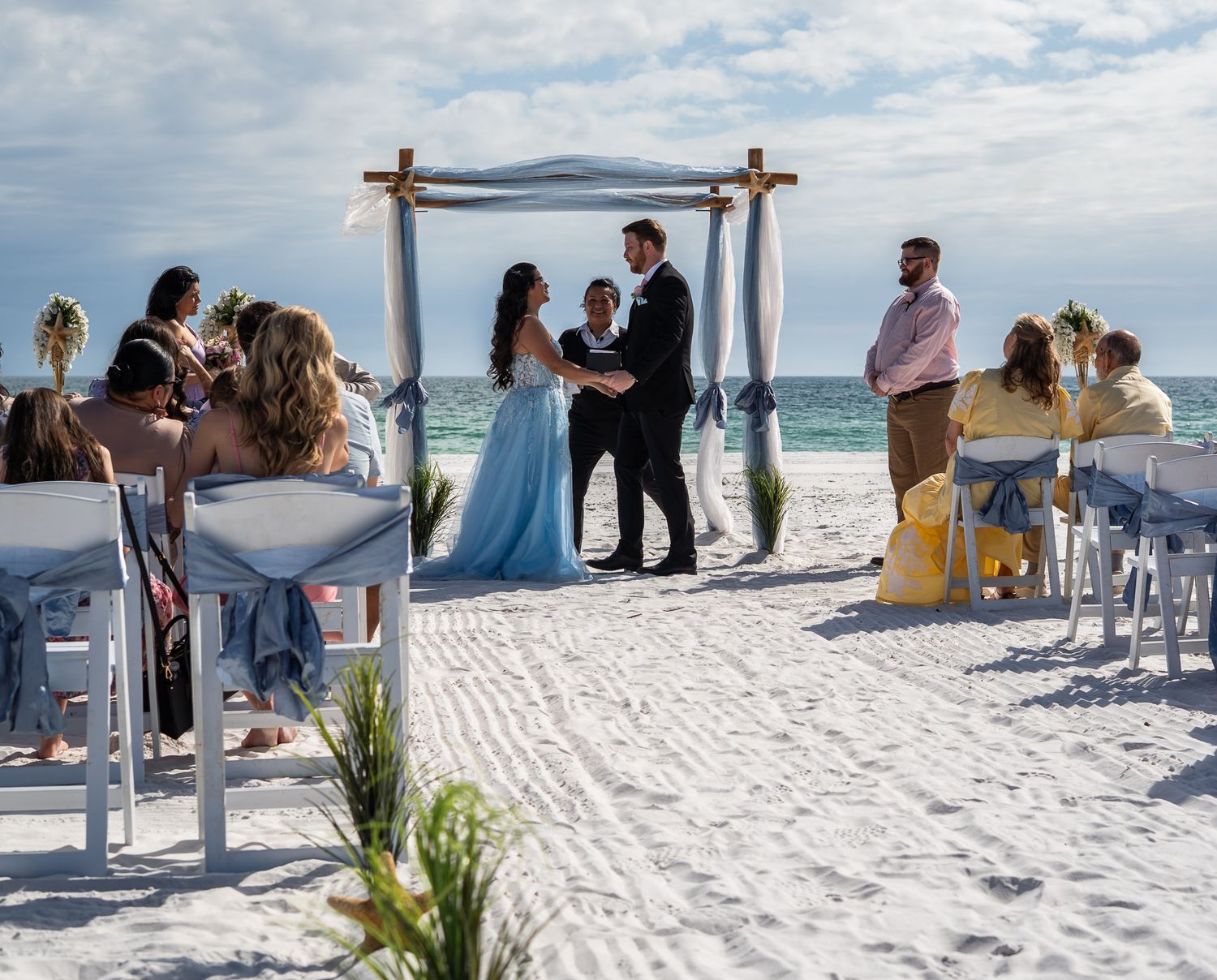 Beach wedding ceremony with bride in light blue gown standing under a rustic wooden arch.