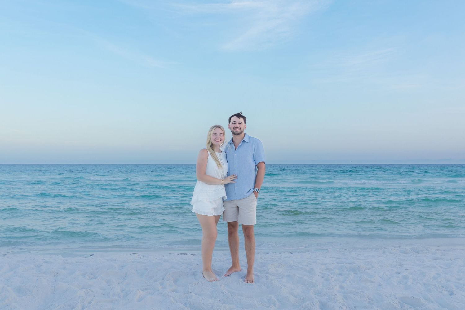 Couple smiling together on a white sand beach with turquoise ocean water and blue sky backdrop.