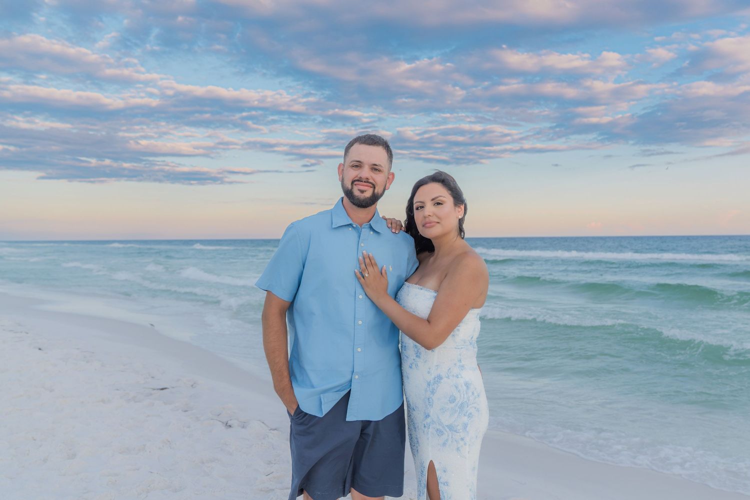 Smiling couple poses together on white sand beach at sunset, man in blue shirt and woman in white dress.