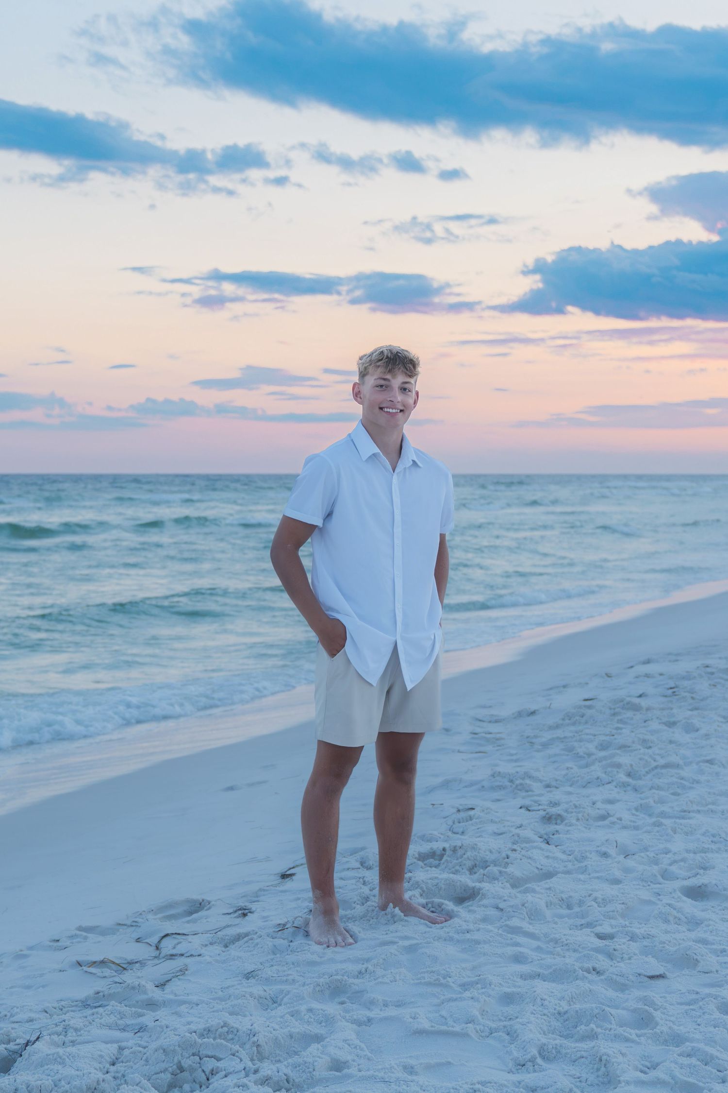 A smiling teenage boy in white shirt and khaki shorts stands barefoot on a sandy beach at sunset.
