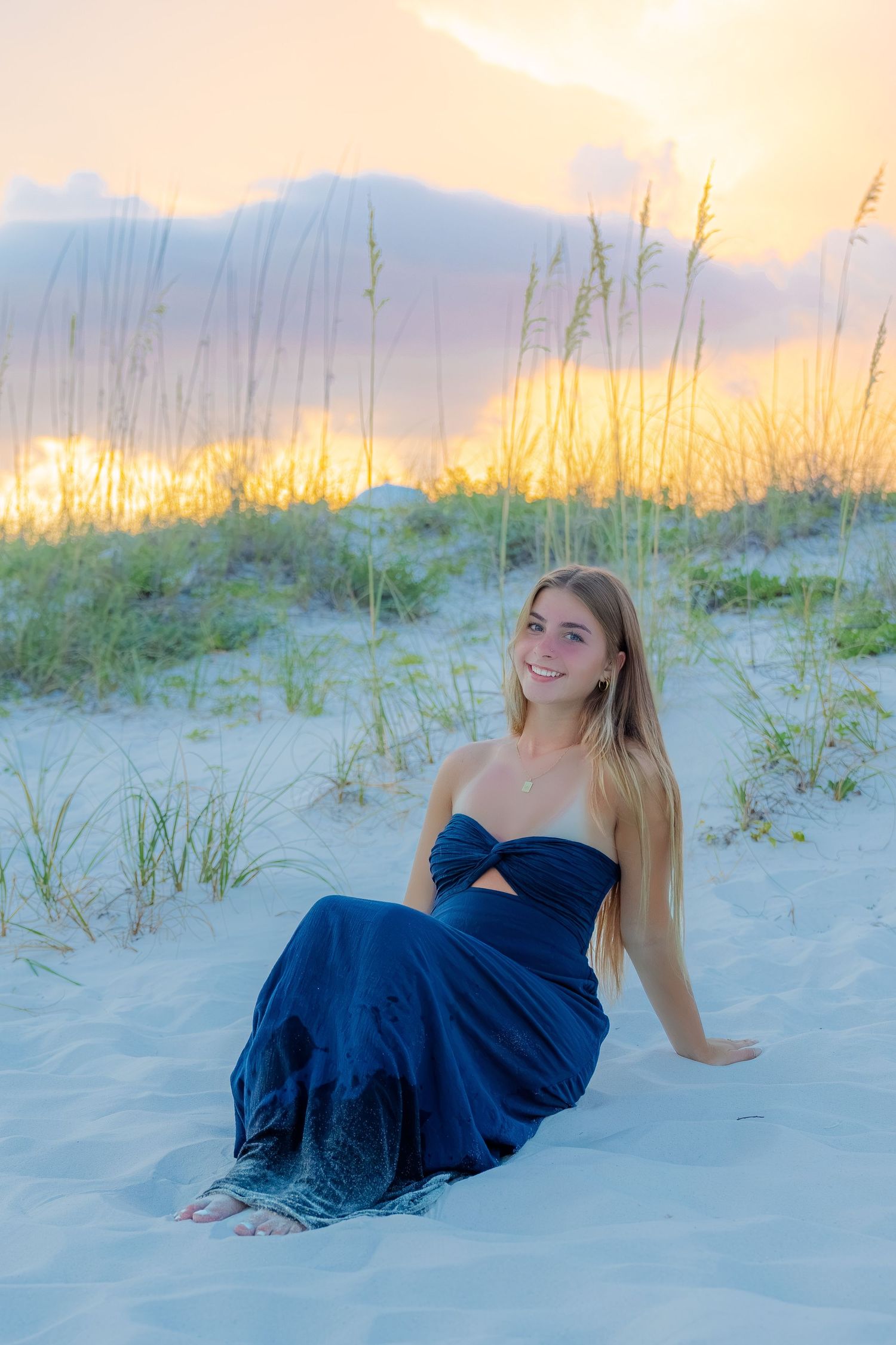 Young blonde woman in a navy blue dress sitting on white sand dunes at sunset with sea oats in background.