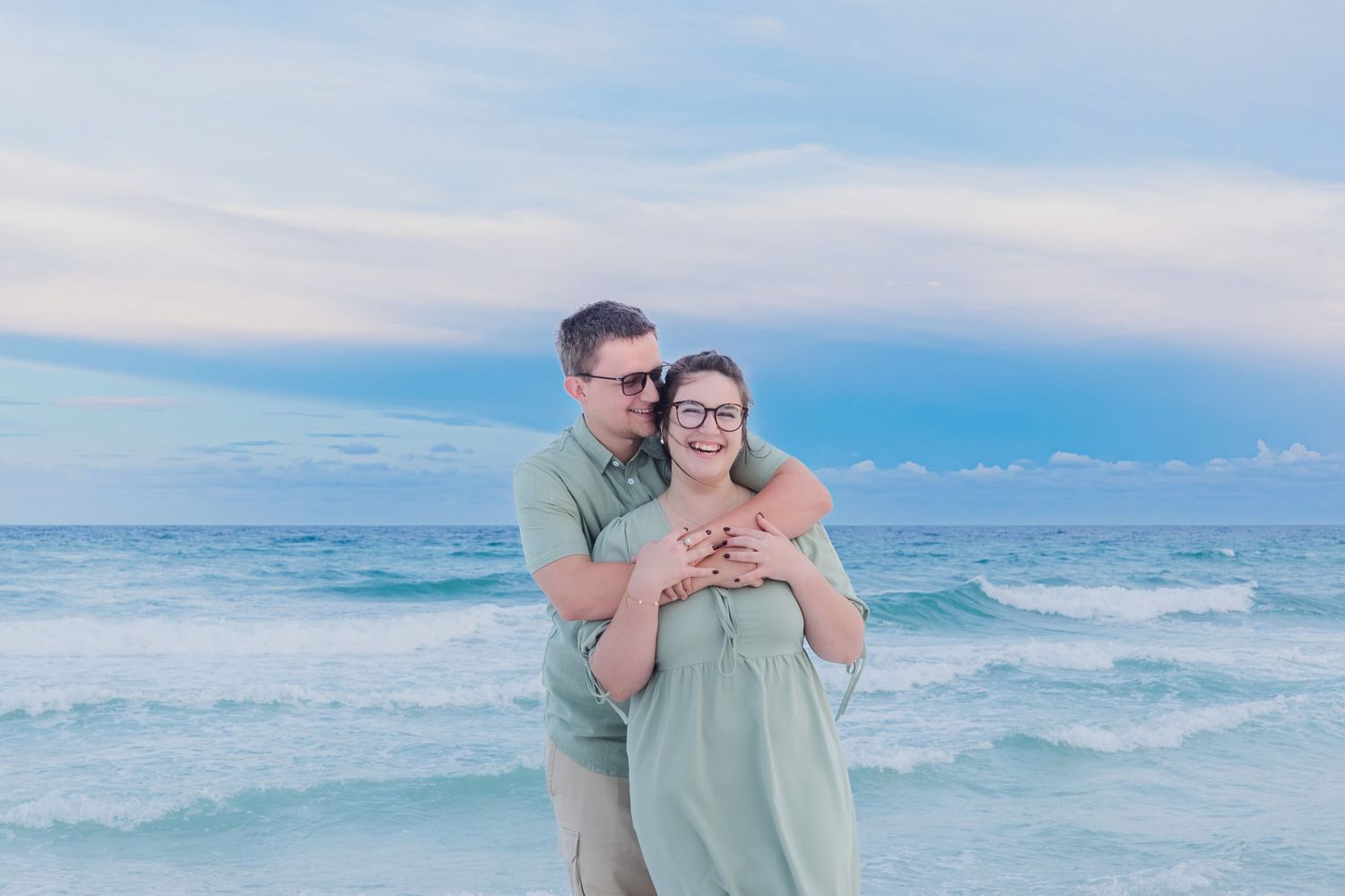 Couple embracing on a sandy beach with turquoise ocean waves and a pastel blue sky at sunset.