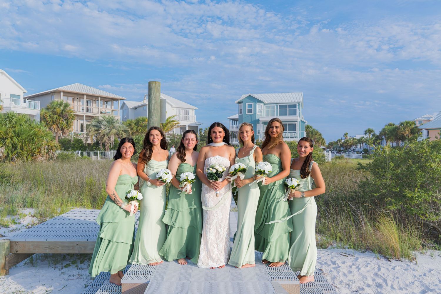Bride and six bridesmaids in mint green dresses pose on a beach boardwalk holding white bouquets.