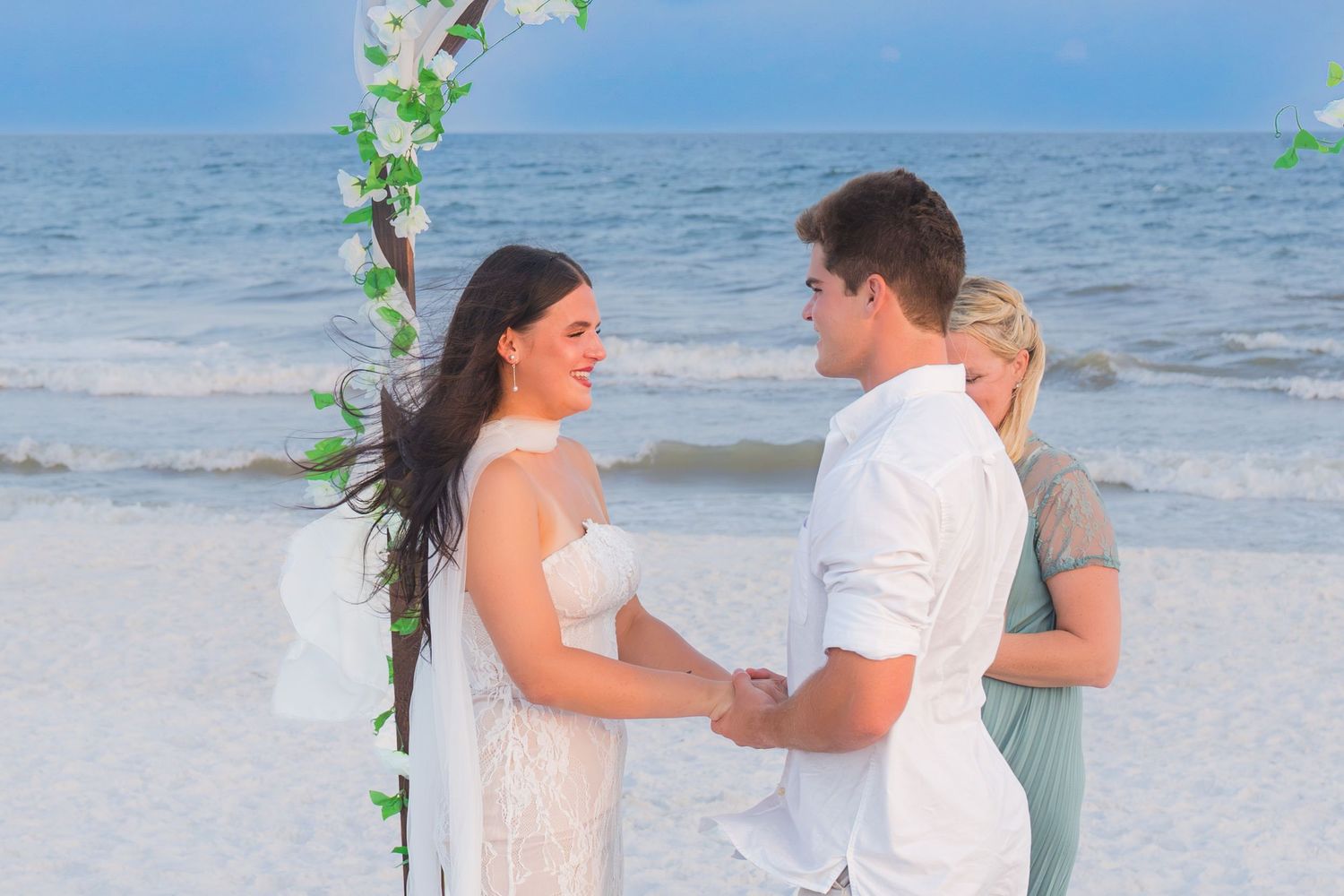 Couple exchanging vows at a beach wedding ceremony with a floral arch and ocean waves in the background.