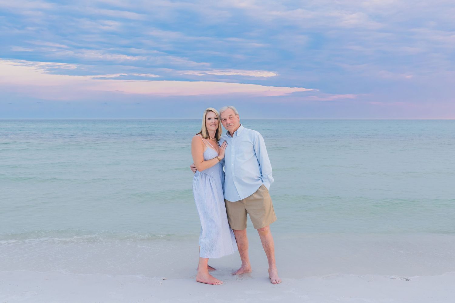 Couple standing barefoot on beach at dusk with turquoise ocean and pastel sky behind them.