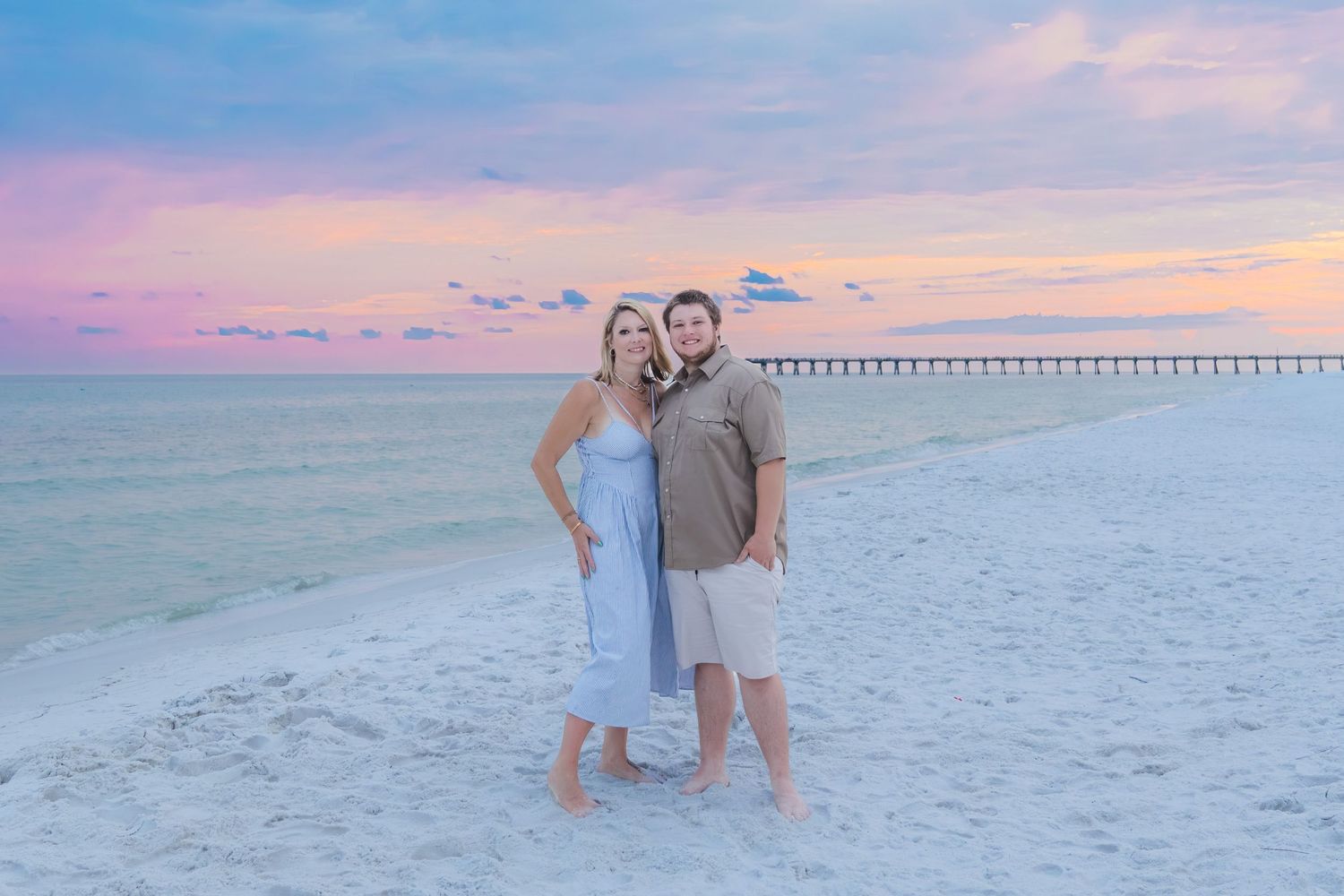 Romantic couple posing on white sand beach at colorful pink and blue sunset near pier.