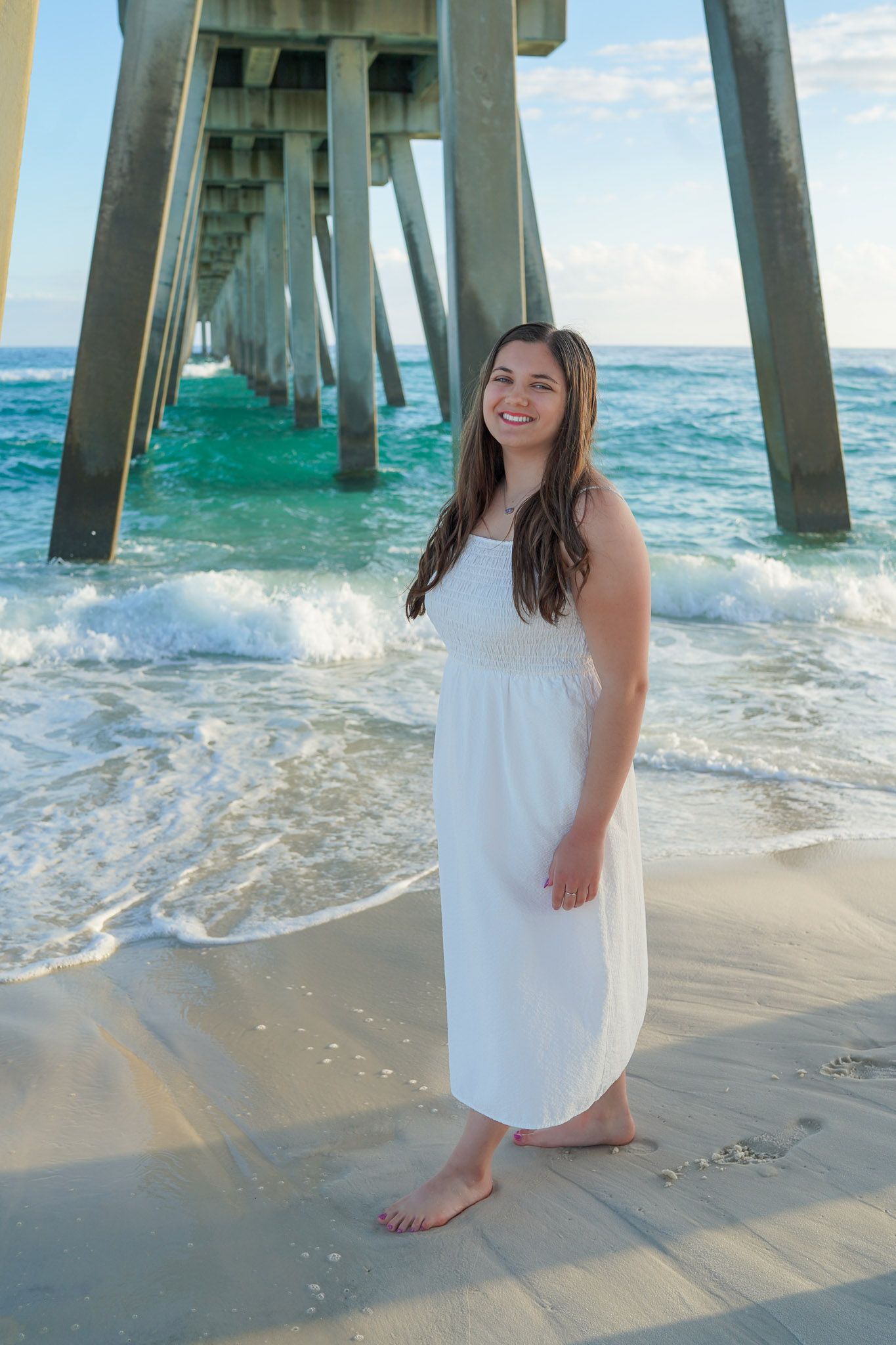 Happy young woman in a white sundress stands barefoot on a beach beside a tall wooden pier.