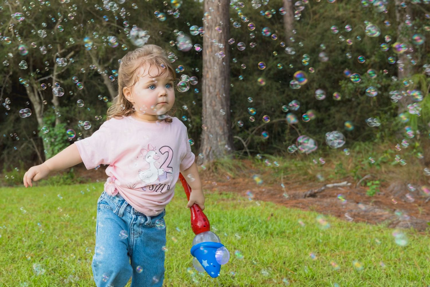 A toddler girl in a pink shirt and jeans plays with a bubble wand outdoors, surrounded by floating bubbles.