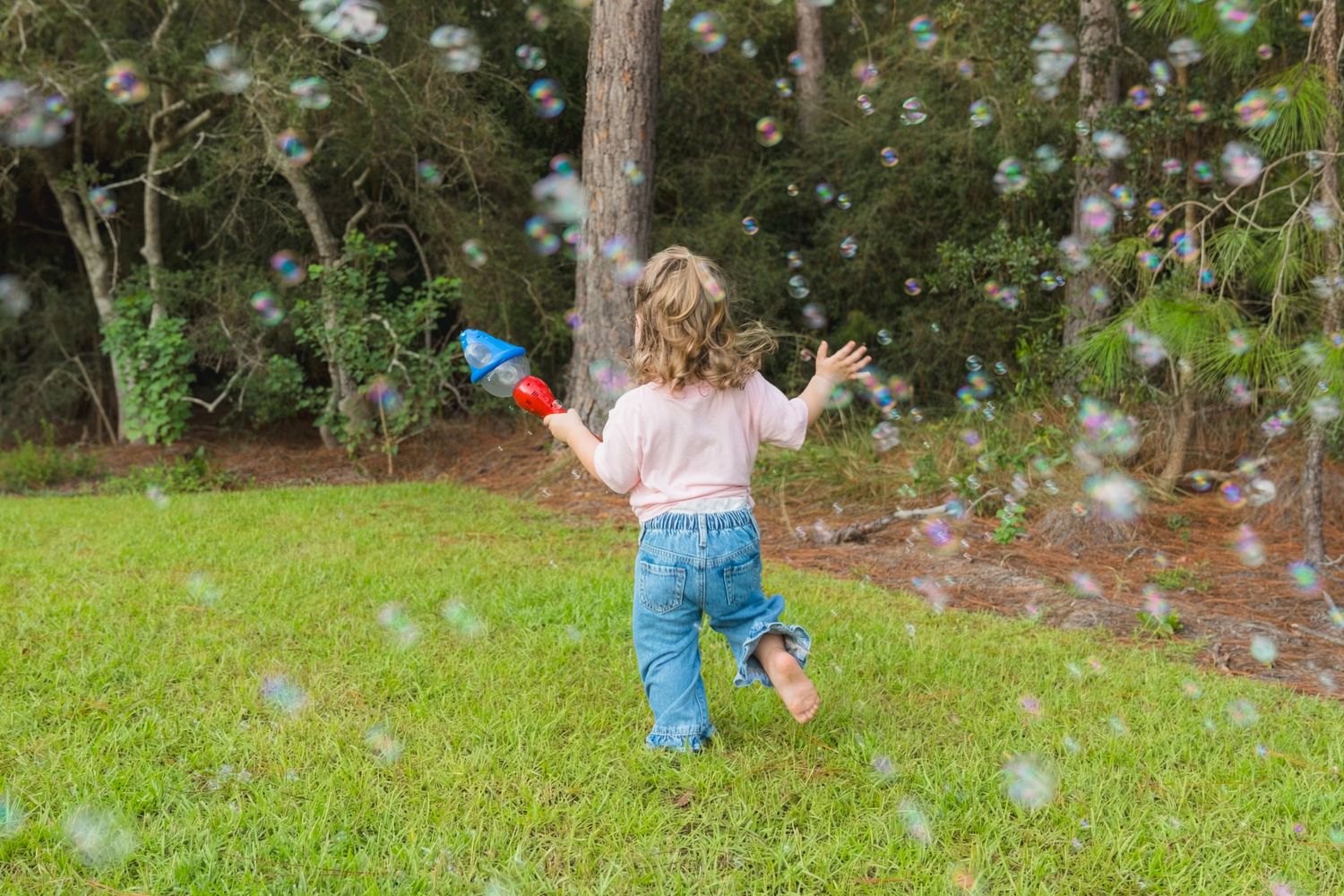 A young child runs through a green yard chasing colorful bubbles while holding a red bubble gun toy.