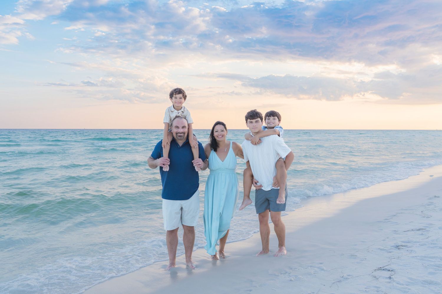 Family of five posing on a sandy beach at sunset, with turquoise water and pastel sky in the background.