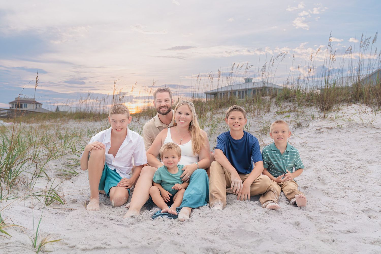 Family of six poses on sandy beach at sunset with sea oats and pastel sky in background.
