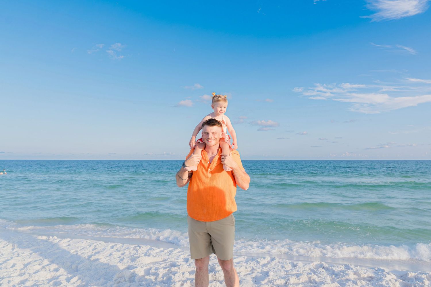 Father carrying his young daughter on his shoulders at a sunny beach with blue ocean waves behind them.