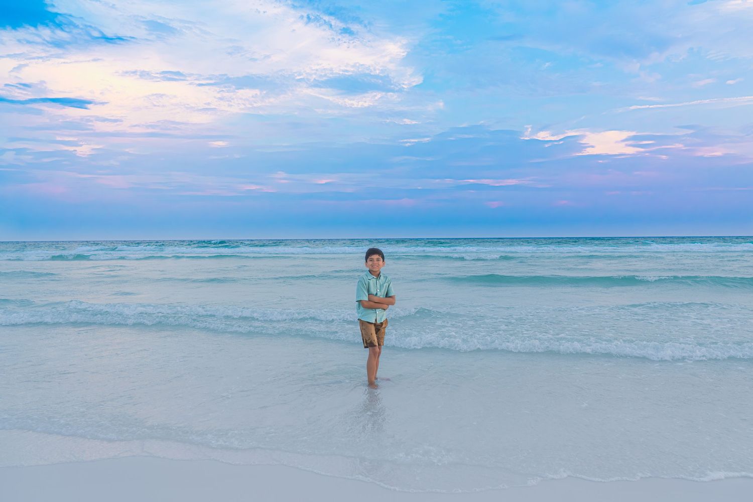 Child standing on a white sand beach at sunset with turquoise waves and a pastel pink and blue sky.