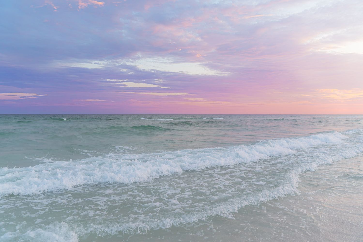 Pastel pink and purple sunset sky over calm turquoise ocean waves gently washing onto a sandy beach.