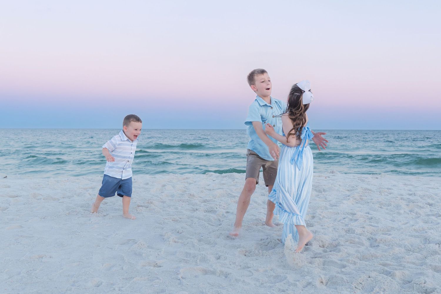 Family of three playing on a sandy beach at sunset, with pastel pink and blue sky over calm ocean waves.