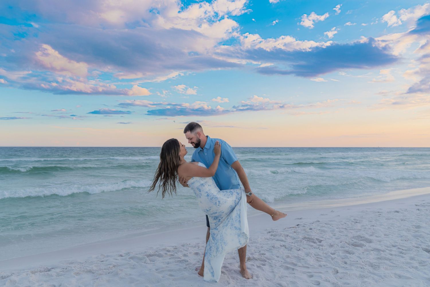Couple sharing a romantic dip kiss on a white sand beach at sunset with turquoise waves and pastel sky.