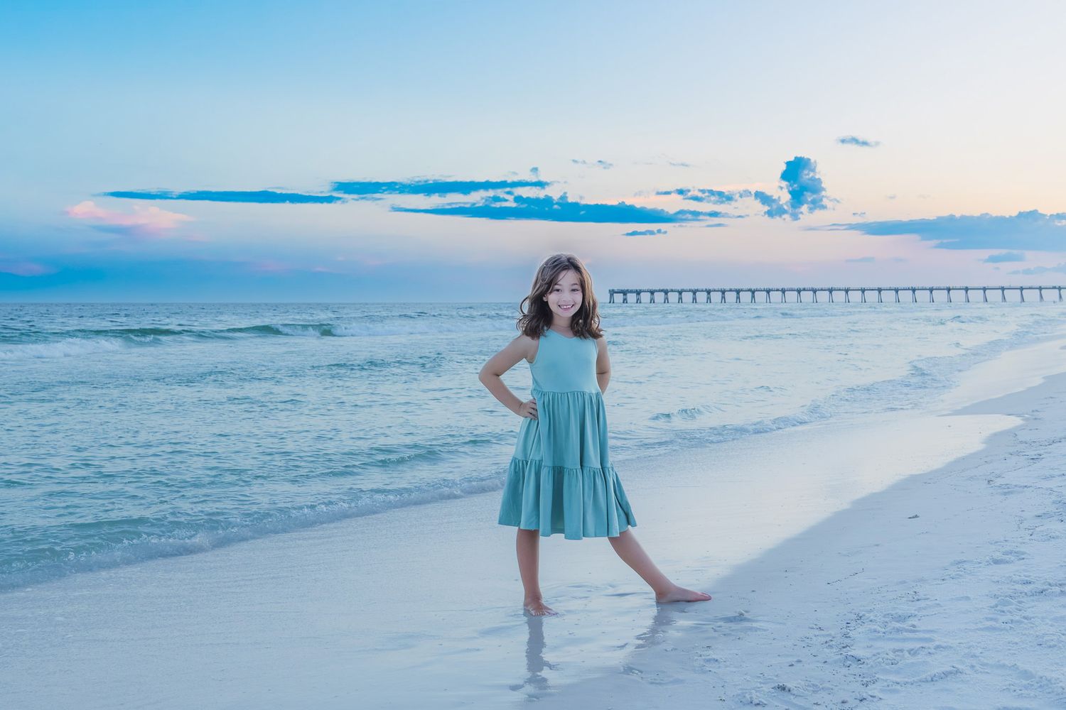 A young girl in a teal dress poses on a sandy beach at sunset with a pier visible in the background.
