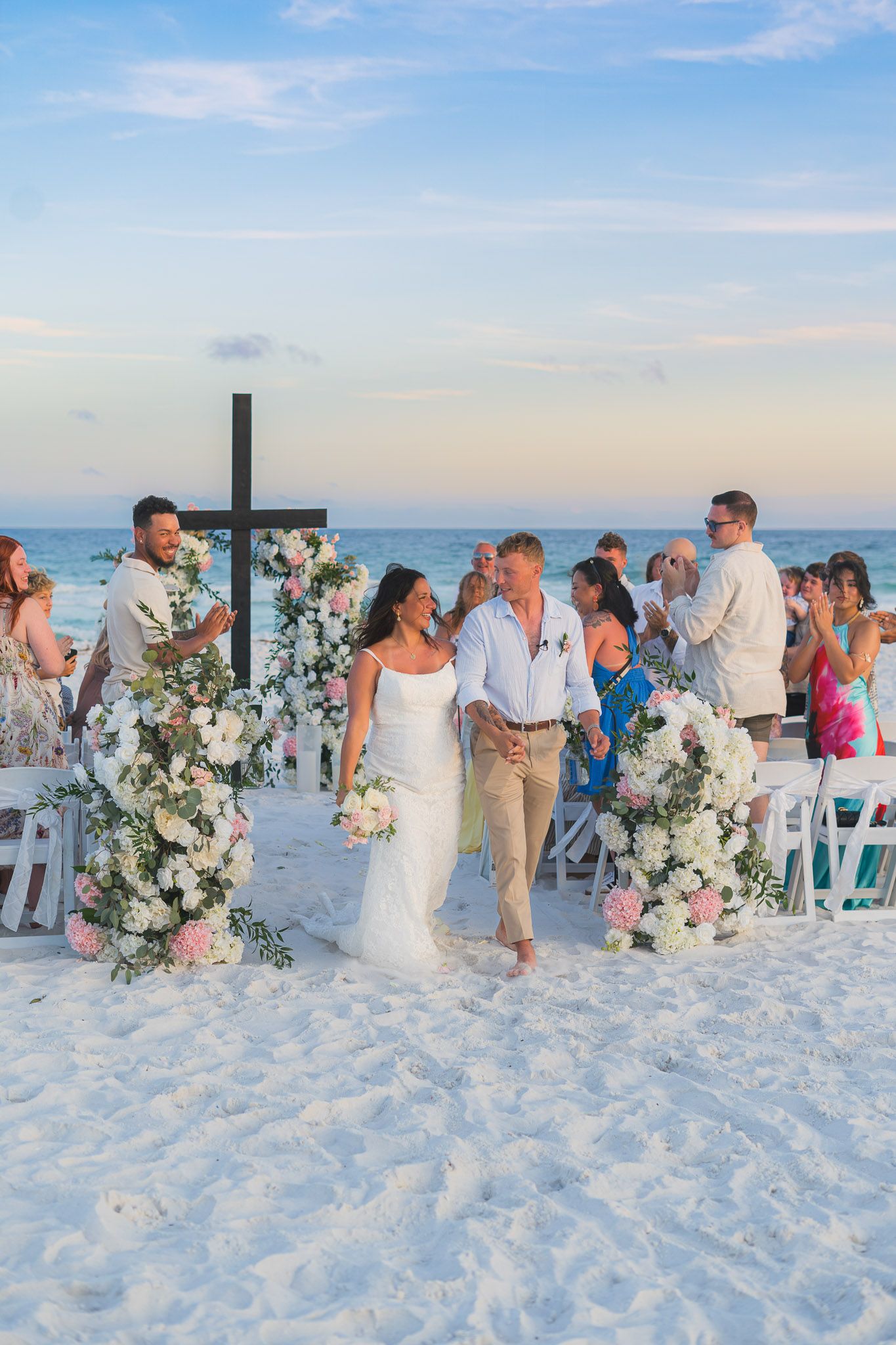 Bride and groom walk down sandy beach aisle flanked by floral arches with white and pink blooms at sunset ceremony.